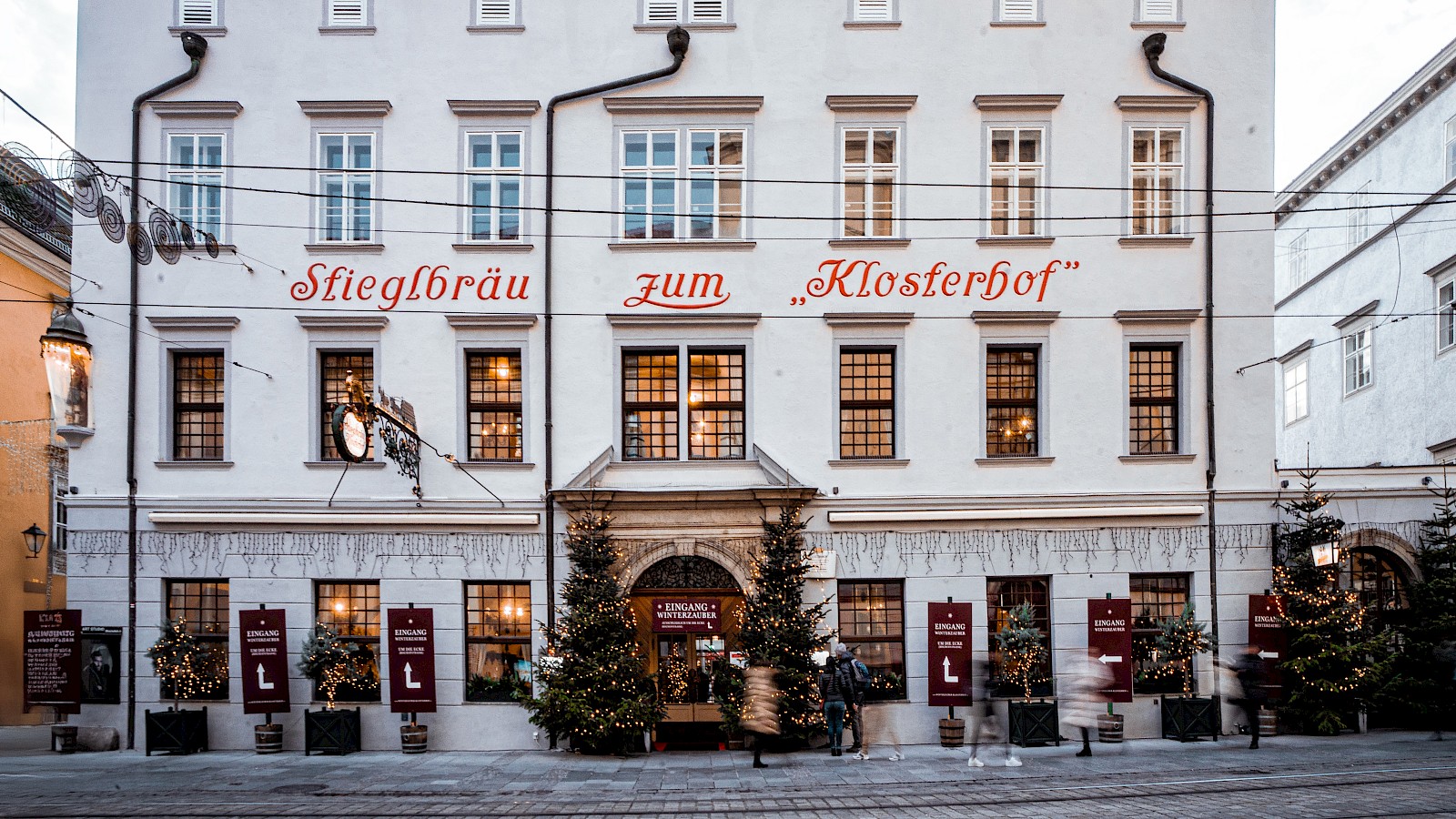 Das Bild zeigt die Außenansicht des Traditionsgasthauses „Stieglbräu zum Klosterhof“ in der Linzer Altstadt. Die historische Fassade mit ihren hellen Wänden und großen Fenstern vermittelt klassischen Charme. Auf dem Gehsteig davor sind gemütliche Holztische und -stühle für den Schanigarten aufgestellt, ideal für Gäste, die bei schönem Wetter draußen speisen möchten. Über dem Eingang prangt ein kunstvolles Wirtshausschild. Das Gebäude befindet sich an einer belebten Straße mit Straßenbahnschienen und ist von weiteren Altbauten gesäumt – ein einladendes Ambiente für traditionelle Gastlichkeit mitten in der Stadt.