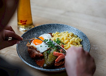 Das Bild zeigt ein köstlich gebratenes ganzes Hähnchen, das auf einem weißen Teller angerichtet ist. Die Haut ist goldbraun und knusprig, was auf eine perfekte Zubereitung hinweist. Auf dem Hähnchen liegt ein Zweig frischer Rosmarin, der nicht nur dekorativ wirkt, sondern dem Gericht auch ein würziges Aroma verleiht. Daneben steht ein kleiner Metallbehälter, vermutlich mit einer passenden Soße oder Dip. Der Hintergrund ist unscharf, aber man erkennt rote und schwarze Farbtöne, was dem Bild eine warme, gemütliche Atmosphäre verleiht – ideal für ein traditionelles, herzhaftes Essen.