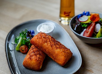 Erdäpfel Cordon Bleu (c) Gassner Gastronomie, Foto Hechtfisch: Zwei goldbraun gebackene Spezialitäten liegen auf einem modernen grauen Teller und wirken knusprig und frisch zubereitet. Dazu werden ein kleiner Dip sowie frischer Salat mit Kräutern, Tomaten und essbaren Blüten serviert. Im Hintergrund steht eine Schüssel mit buntem Blattsalat und frischem Gemüse. Ein Glas helles Bier ergänzt das Gericht und sorgt für typisch österreichischen Genuss. Die warme Beleuchtung und der schlichte Holztisch schaffen eine gemütliche und stilvolle Atmosphäre. Gäste erleben hier hochwertige Küche in entspannter und einladender Umgebung.