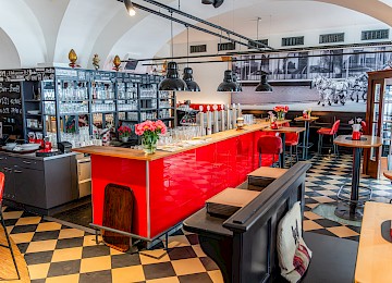Das Bild zeigt einen Innenraum mit charakteristischen weißen Gewölbedecken und modernen Elementen. Die Deckenbögen verleihen dem Raum eine historische, fast kirchliche Atmosphäre, während moderne Lichtinstallationen – lange schwarze Kabel mit schlichten Hängelampen – einen stilvollen Kontrast setzen. Die Wände sind weiß gestrichen und reflektieren das Licht sanft, wodurch der Raum offen und freundlich wirkt. Der Gesamteindruck ist eine gelungene Mischung aus traditioneller Architektur und zeitgenössischem Design. Dieser Bereich könnte Teil eines gehobenen Restaurants oder eines stilvoll renovierten Altbaus sein, der Geschichte und Moderne harmonisch verbindet.