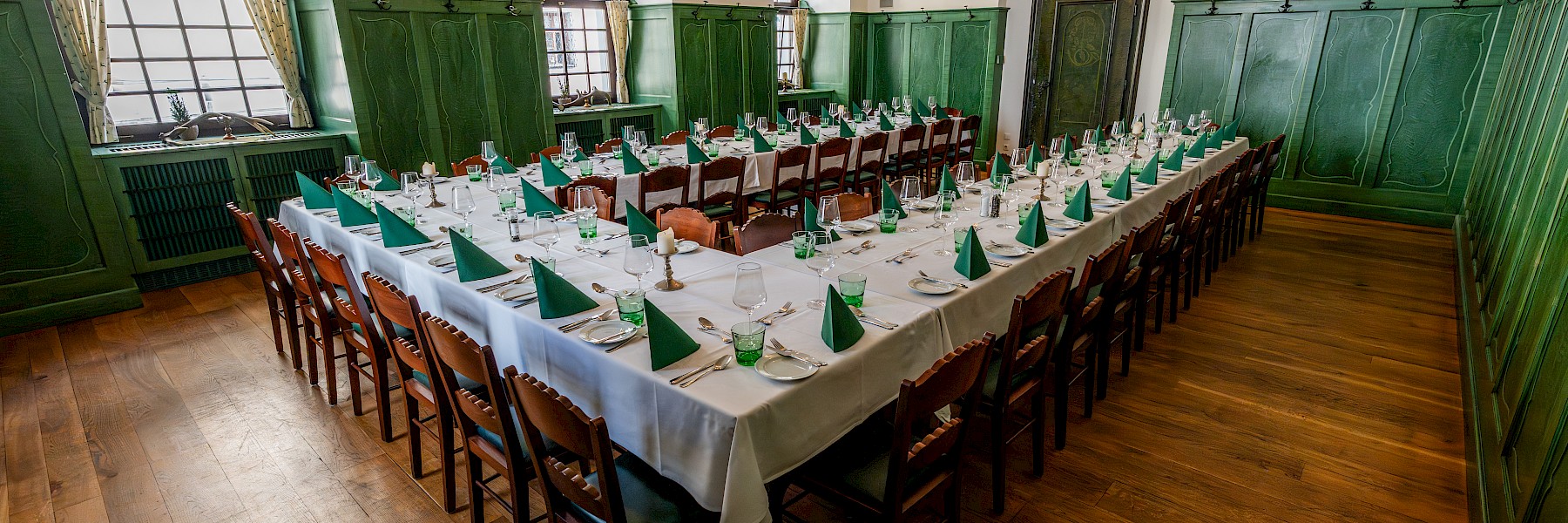 Braustube (c) Gassner Gastronomie: Das Bild zeigt einen festlich und traditionell eingerichteten Gastraum im Klosterhof mit warmem, stimmungsvollem Licht. Die Tische sind mit weißen Tischläufern und silbernen Tabletts mit Kerzen dekoriert, die eine gemütliche Atmosphäre schaffen. Die dunklen Holzmöbel stehen im Kontrast zu den hellen Wänden und dem kunstvoll verzierten Wandbild über einer alten Holztür. Große Fenster mit hellen Vorhängen lassen Tageslicht hinein, während weihnachtliche Kränze und kleine Dekorationen für saisonales Flair sorgen. Der Raum vermittelt eine elegante, alpenländische Gemütlichkeit, ideal für gesellige Runden in stilvollem Ambiente.