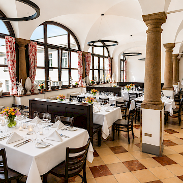 Aula (c) Gassner Gastronomie: Die „Aula“ wirkt hell, großzügig und modern zugleich. Große Rundbogenfenster lassen viel Tageslicht in den Raum und schaffen eine freundliche Atmosphäre. Hohe steinerne Säulen verleihen dem Saal Eleganz und historischen Charakter. Helle Böden und schlicht gedeckte Holztische sorgen für ein gepflegtes und ruhiges Ambiente. Dunkle Stühle bieten gemütliche Sitzplätze für Gäste. Moderne runde Deckenlampen setzen stilvolle Akzente und ergänzen die historische Architektur. Die großzügige Raumaufteilung eignet sich ideal für gemeinsame Essen, Veranstaltungen oder entspannte Stunden in angenehmer Umgebung.
