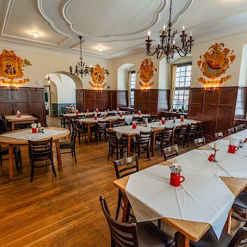 Wappenzimmer (c) Gassner Gastronomie: Das „Wappenzimmer“ bietet eine traditionelle und elegante Atmosphäre mit historischem Charakter. Dunkle Holzvertäfelungen und große Holztische verleihen dem Raum Wärme und Gemütlichkeit. Historische Wappen an den Wänden geben dem Saal seinen besonderen Namen und unterstreichen die österreichische Tradition. Große Fenster lassen viel Tageslicht herein und schaffen eine helle, angenehme Stimmung. Klassische Kronleuchter sorgen für stilvolle Beleuchtung und ein festliches Ambiente. Die großzügige Raumaufteilung eignet sich ideal für Familienfeiern, Gruppenessen oder Veranstaltungen.