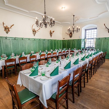 Jagdzimmer (c) Gassner Gastronomie: Das „Jagdzimmer“ verbindet traditionelle österreichische Gemütlichkeit mit historischem Jagdcharme. Die grünen Holzvertäfelungen verleihen dem Raum eine besondere und elegante Atmosphäre. An den Wänden hängen dekorative Geweihe, die den klassischen Stil des Jagdzimmers unterstreichen. Dunkle Holztische und passende Stühle bieten gemütliche Sitzplätze für Gäste. Große Kronleuchter sorgen für warmes Licht und ein stilvolles Ambiente. Der helle Raum wirkt ruhig, gepflegt und einladend. Die großzügige Anordnung der Tische eignet sich ideal für gemeinsame Essen, Feiern oder entspannte Stunden in traditioneller Umgebung.