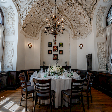 Prälatenstübl (c) Gassner Gastronomie: Das „Prälatenstübl“ beeindruckt mit kunstvoller Architektur und stilvoller Eleganz. Die hohe, reich verzierte Stuckdecke mit floralen Ornamenten verleiht dem Raum eine besondere historische Atmosphäre. Ein großer Kronleuchter sorgt für warmes Licht und unterstreicht das edle Ambiente. In der Mitte steht ein runder Tisch mit weißer Tischdecke und bequemen gepolsterten Stühlen. Dunkle Holzelemente bilden einen eleganten Kontrast zu den hellen Wänden. Kleine Kunstwerke und dekorative Details ergänzen die hochwertige Einrichtung. Der Raum eignet sich ideal für exklusive Essen, private Feiern oder besondere Veranstaltungen in ruhiger und stilvoller Umgebung.