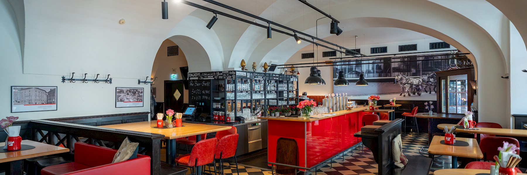 Stieglitz (c) Gassner Gastronomie: The image shows a modern bar – the Stieglitz – with stylish furnishings set in a room with historic vaulted ceilings. In the foreground, a long counter with a bright red front, tall bar stools, and several beer taps takes center stage. Above, simple black pendant lights hang from the ceiling. Behind the bar is a shelving unit filled with glasses and bottles. The floor features a classic black-and-white checkerboard tile pattern. In the background, additional tables and seating areas invite guests to relax. The blend of traditional architecture and modern bar aesthetics creates a welcoming and elegant atmosphere.