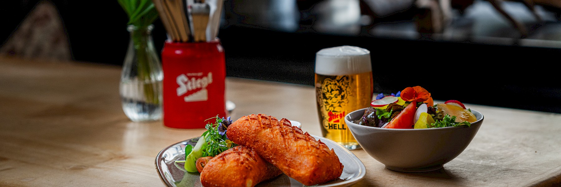Erdäpfel Cordon Bleu (c) Gassner Gastronomie, Foto Hechtfisch: Auf dem Holztisch stehen zwei goldbraun gebackene Speisen auf einem Teller, liebevoll angerichtet mit frischem Salat und Kräutern. Daneben befindet sich eine Schüssel mit buntem Blattsalat, Tomaten und essbaren Blüten. Ein Glas helles Bier mit weißer Schaumkrone ergänzt das Gericht. Kleine Blumenvasen und rote Behälter mit Besteck sorgen für gemütliche Farbakzente. Die warme Beleuchtung und die ruhige Umgebung schaffen eine entspannte und einladende Atmosphäre. Gäste genießen hier österreichische Küche in stilvollem und gemütlichem Ambiente.